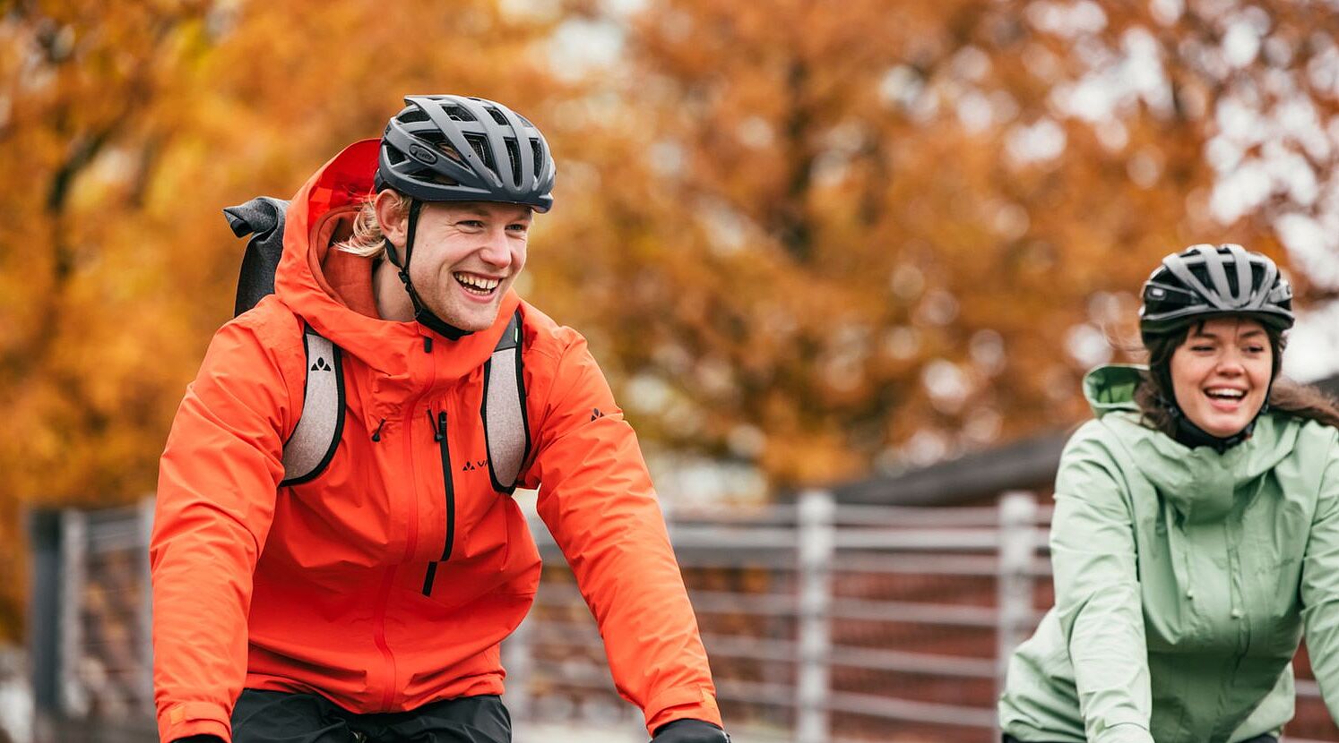 Eine Frau und ein Mann mit Fahrrädern von vorn in herbstlicher Kleidung lächlen in die Kamera. 